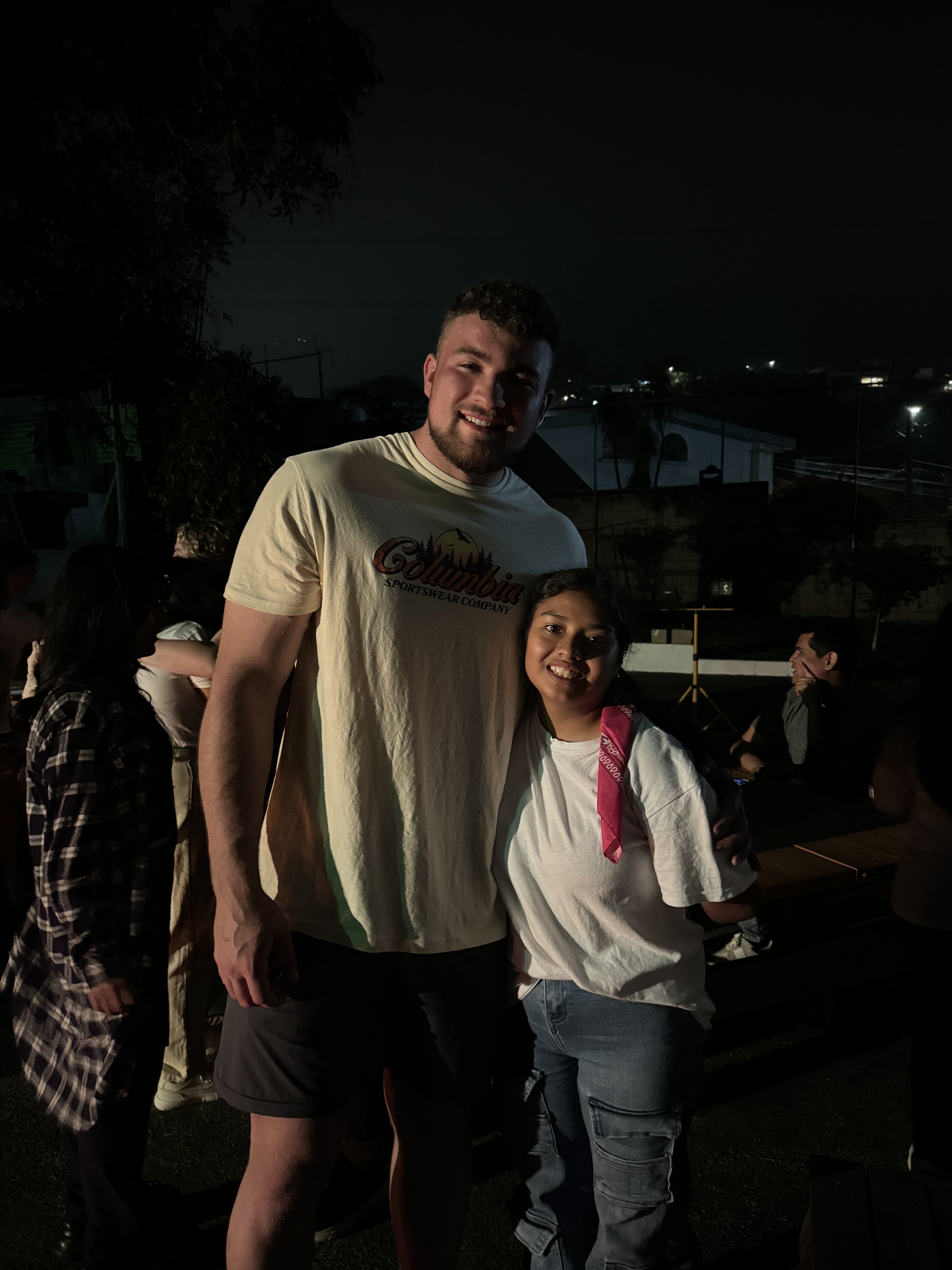 A tall man and a young girl smiling at night outdoors with dark sky and houses in the background.