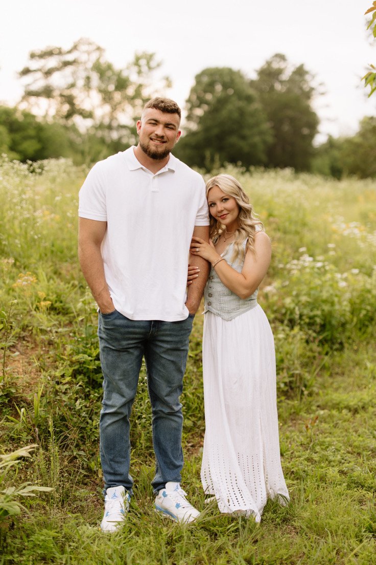 A young couple standing in a grassy field with trees in the background. The man has a beard, short hair, and is wearing a white polo shirt, jeans, and white sneakers. The woman has long blond hair and is wearing a light-colored sleeveless top and a long white skirt. They are smiling and standing close together, with the woman holding onto the man's arm.