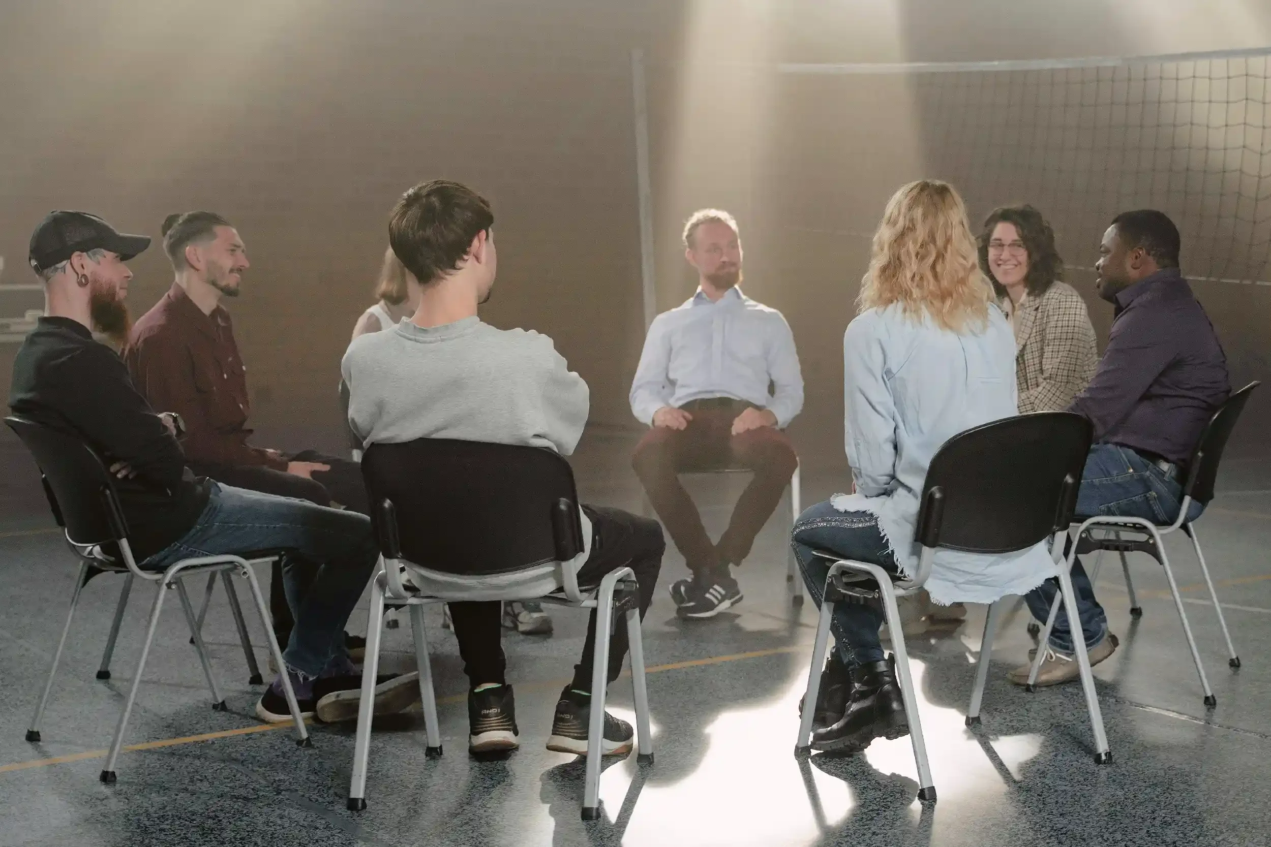 A group of eight diverse people sitting in a circle in a well-lit indoor space, engaging in a discussion.