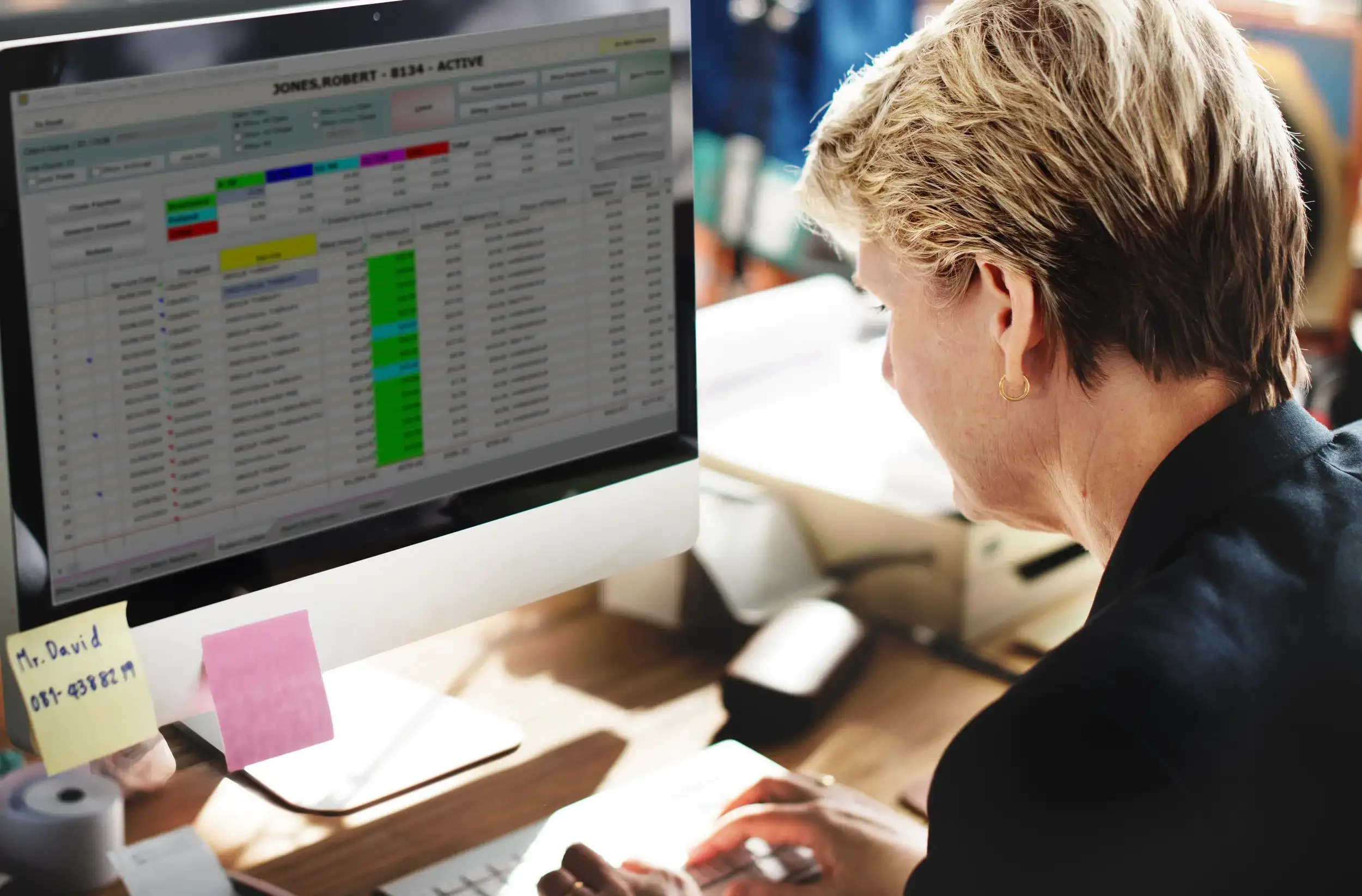 A woman with short blonde hair working at a desk with a desktop computer display showing a spreadsheet with colorful cells and rows.