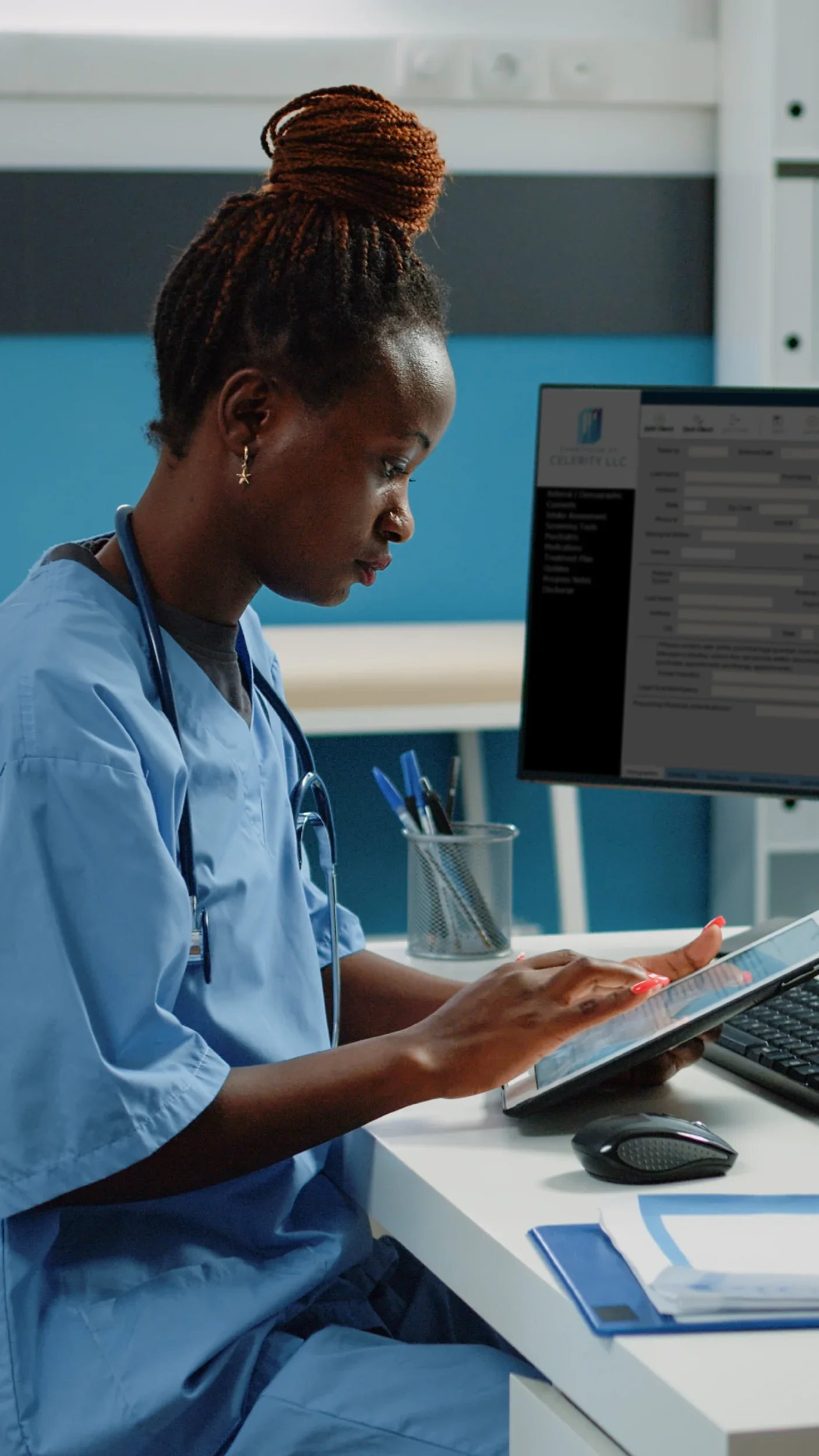 A nurse in blue scrubs sitting at a white desk, looking at a digital tablet. There is a computer monitor, a keyboard, a mouse, and a pen holder on the desk.