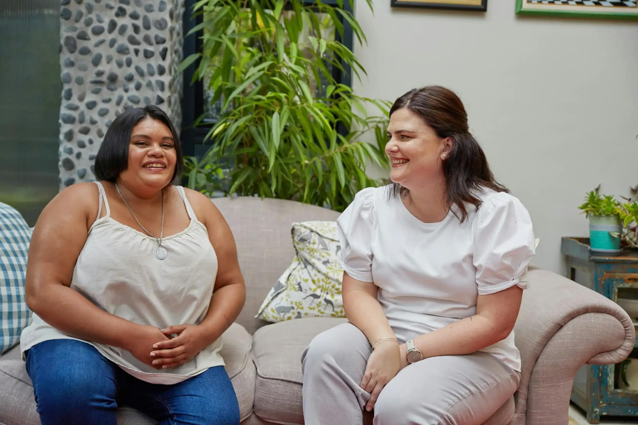 Two women sitting on a beige couch, talking and smiling in a living room with green plants and framed pictures on the wall.