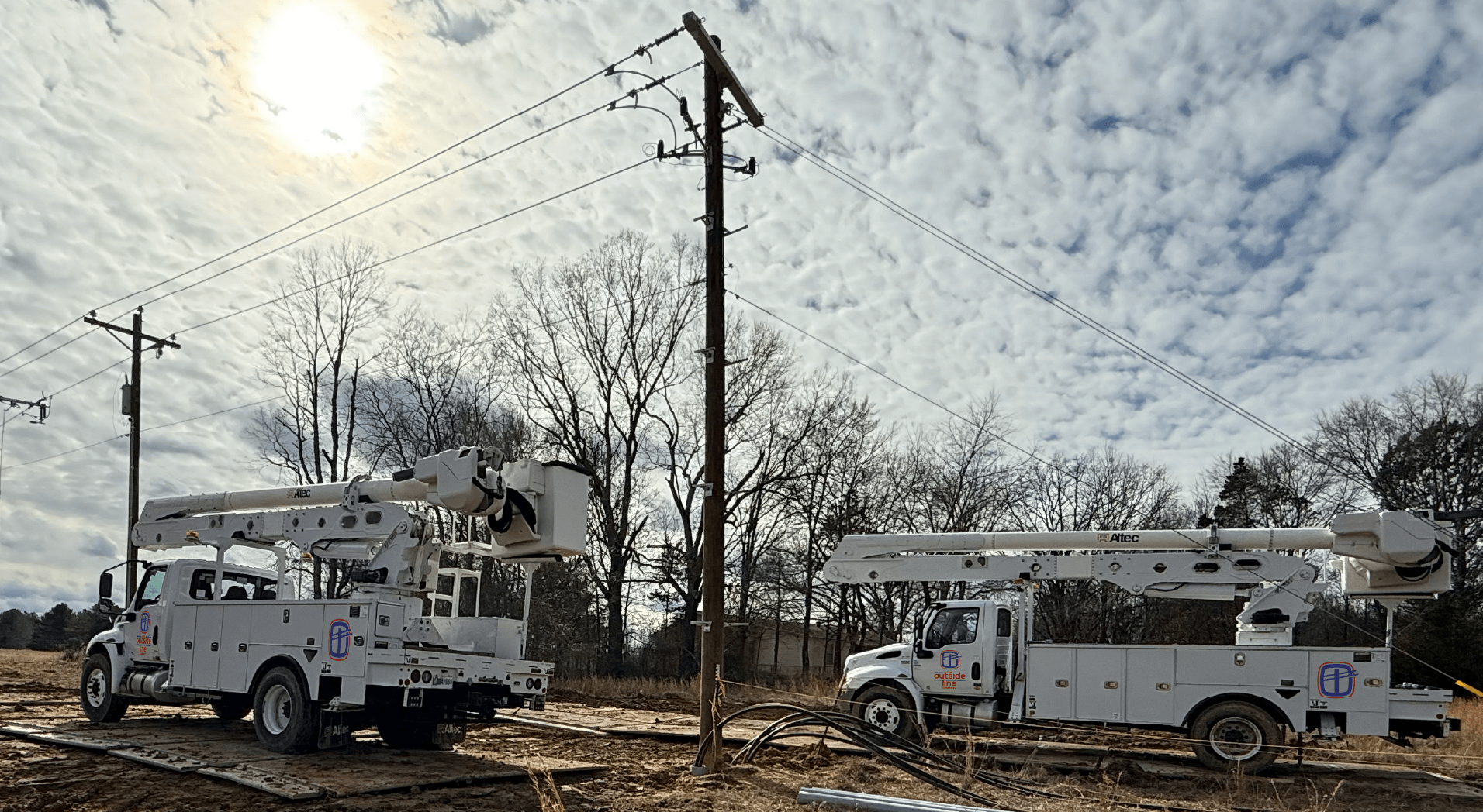 Two utility bucket trucks working on power lines at a construction site with dirt ground and leafless trees in the background, under a cloudy sky with the sun partially visible.