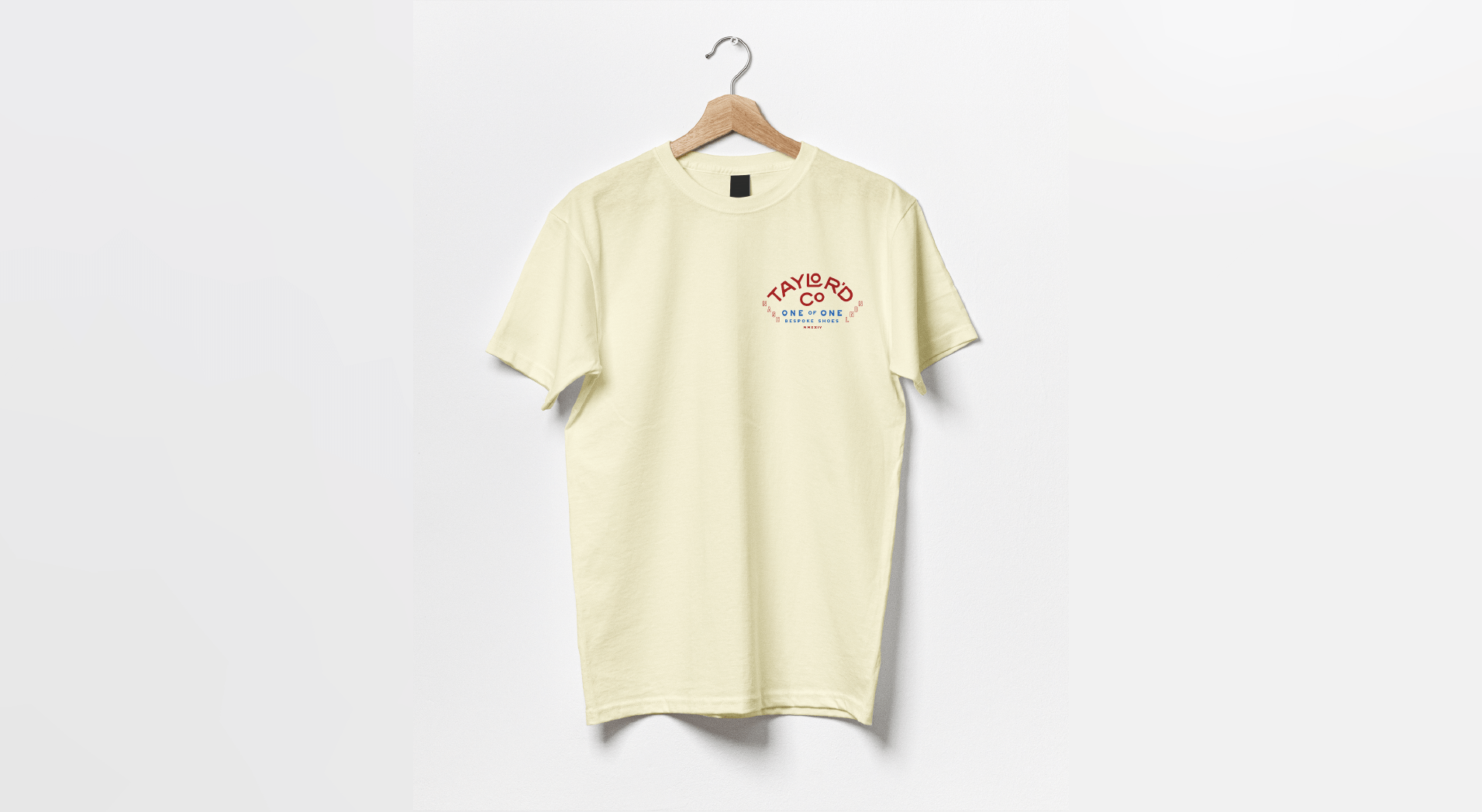 Cream-colored T-shirt hanging on a wooden hanger on a white wall. The shirt has a small red and blue graphic on the upper left chest area that says "TAYLOR'D CO" and "ONE OF ONE" with additional small text.