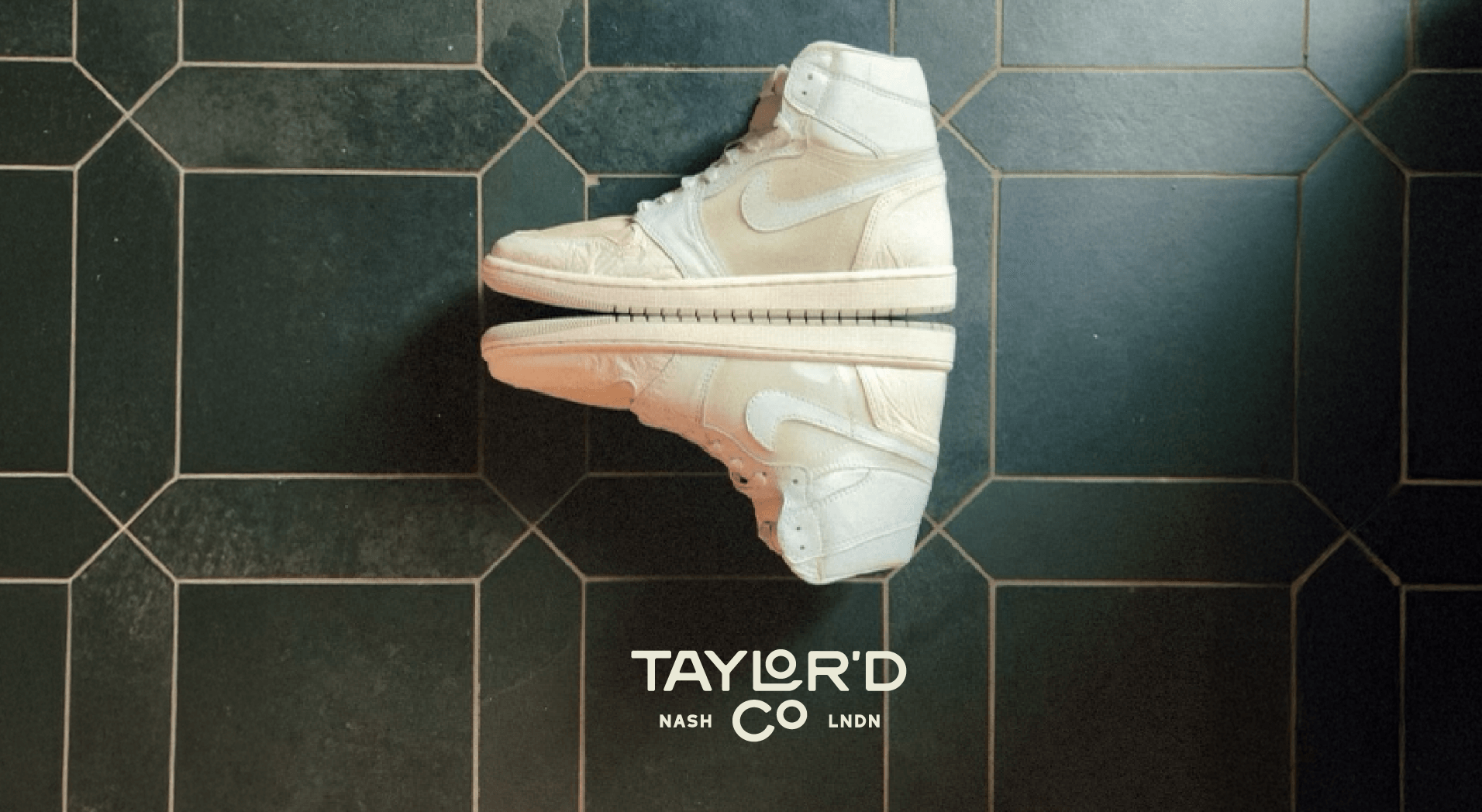 Pair of white high-top sneakers with Nike logo, placed on a black tiled floor with white grout lines. Text overlay reads 'TAYLOR'D CO'.