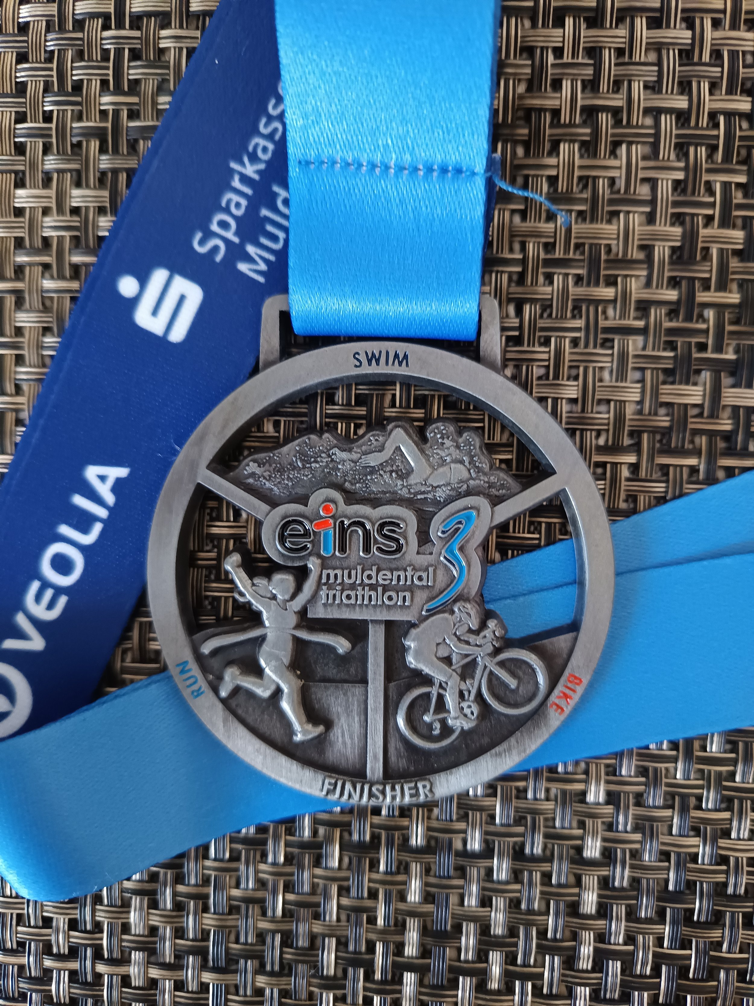 Eine Medaille vom 'ei:ns voll der triathlon 3' mit Abbildungen eines Schwimmers, Radfahrers, Läufers und dem Text 'Swim', 'Bike', 'Run' auf einem braunen Untergrund.