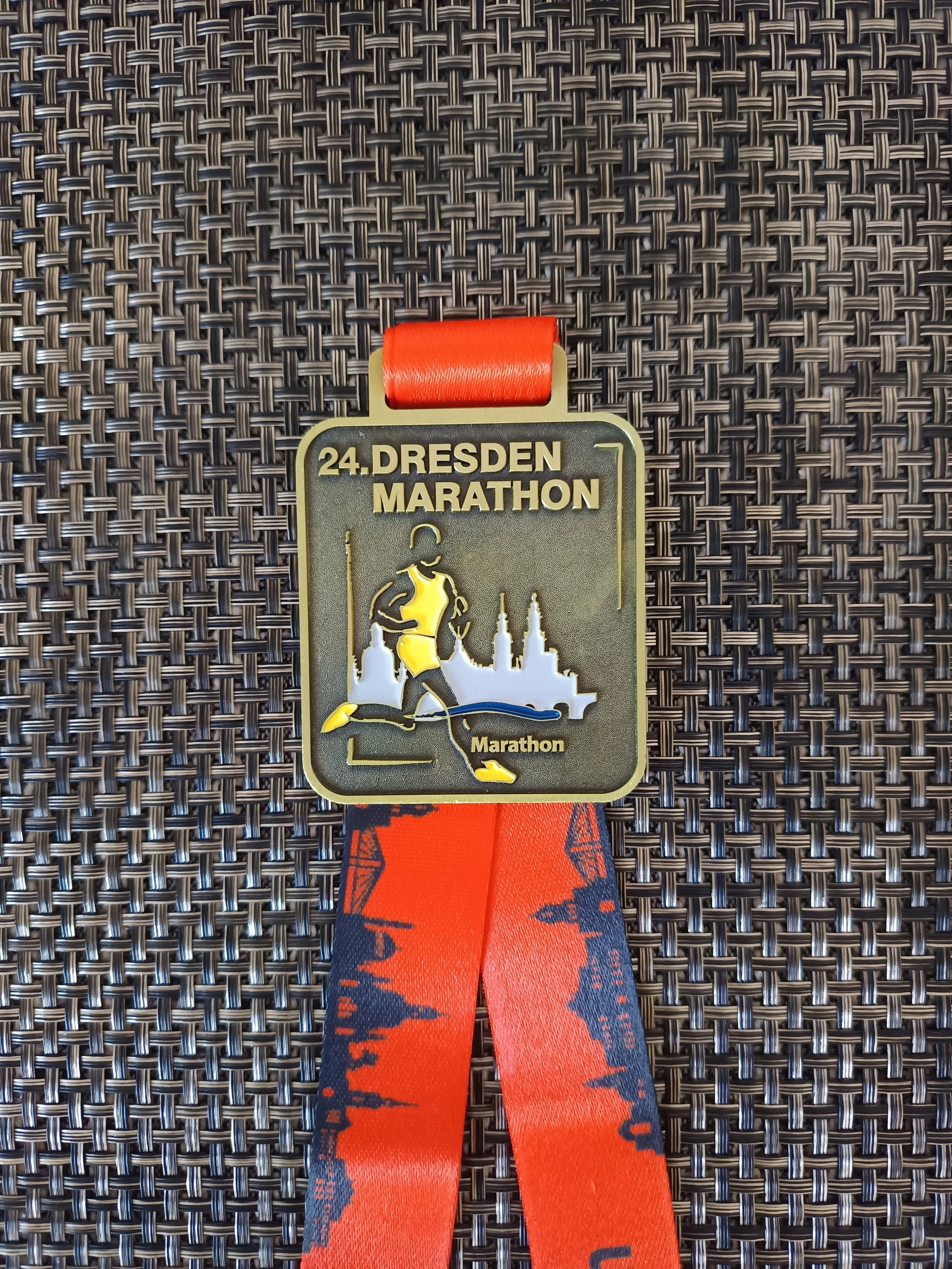 Medaille des 24. Dessau Marathon in Gold und Silber mit orangeem Band, auf braunem Hintergrund.