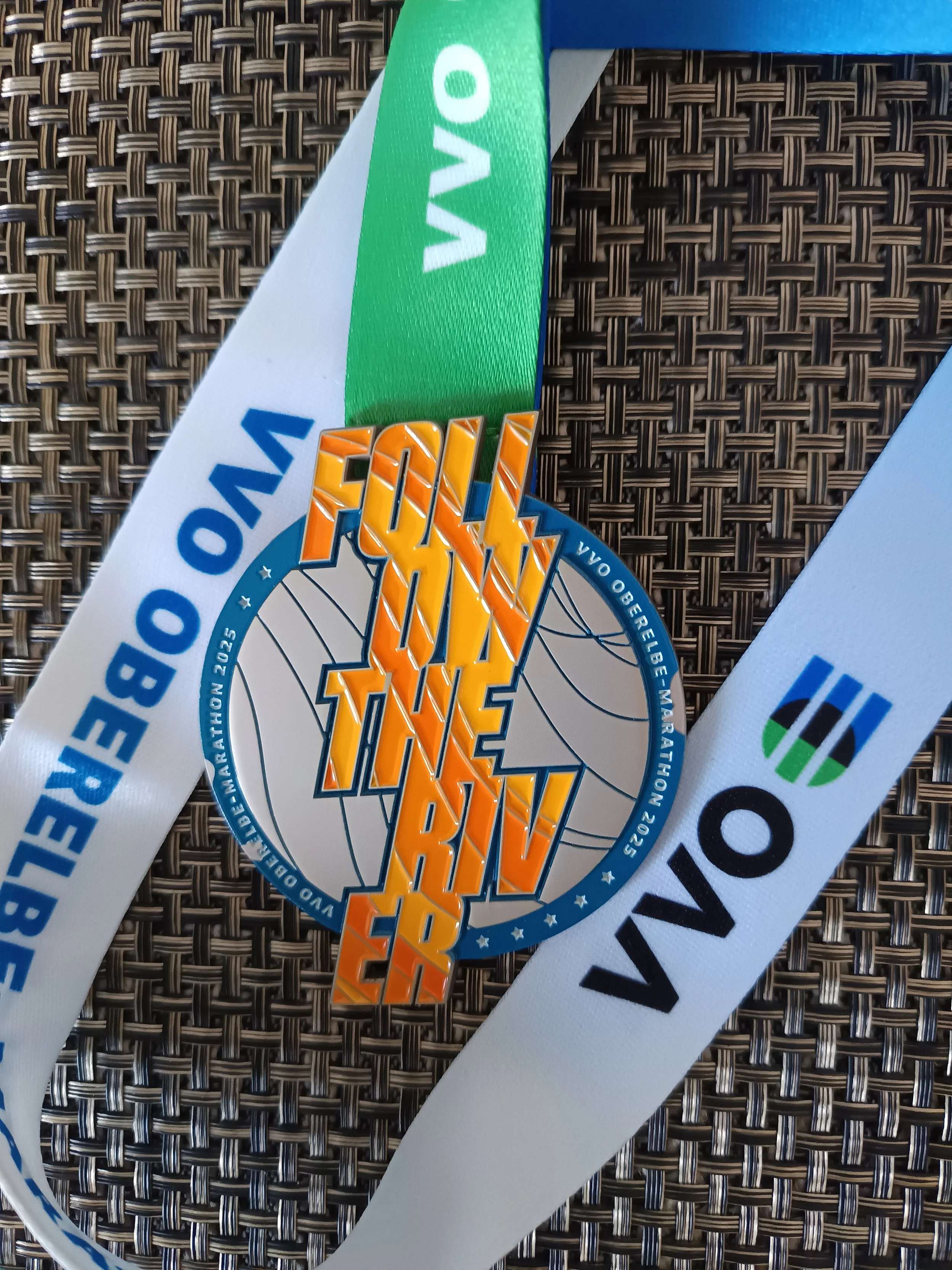 Ein Medaillenträger mit einem blauen Band, das grün, blau und weiß ist, liegt auf einem schwarzen, strukturierten Untergrund. Die Medaille zeigt eine Weltkarte mit dem Schriftzug 'FINISHER' in großen orangefarbenen Buchstaben.