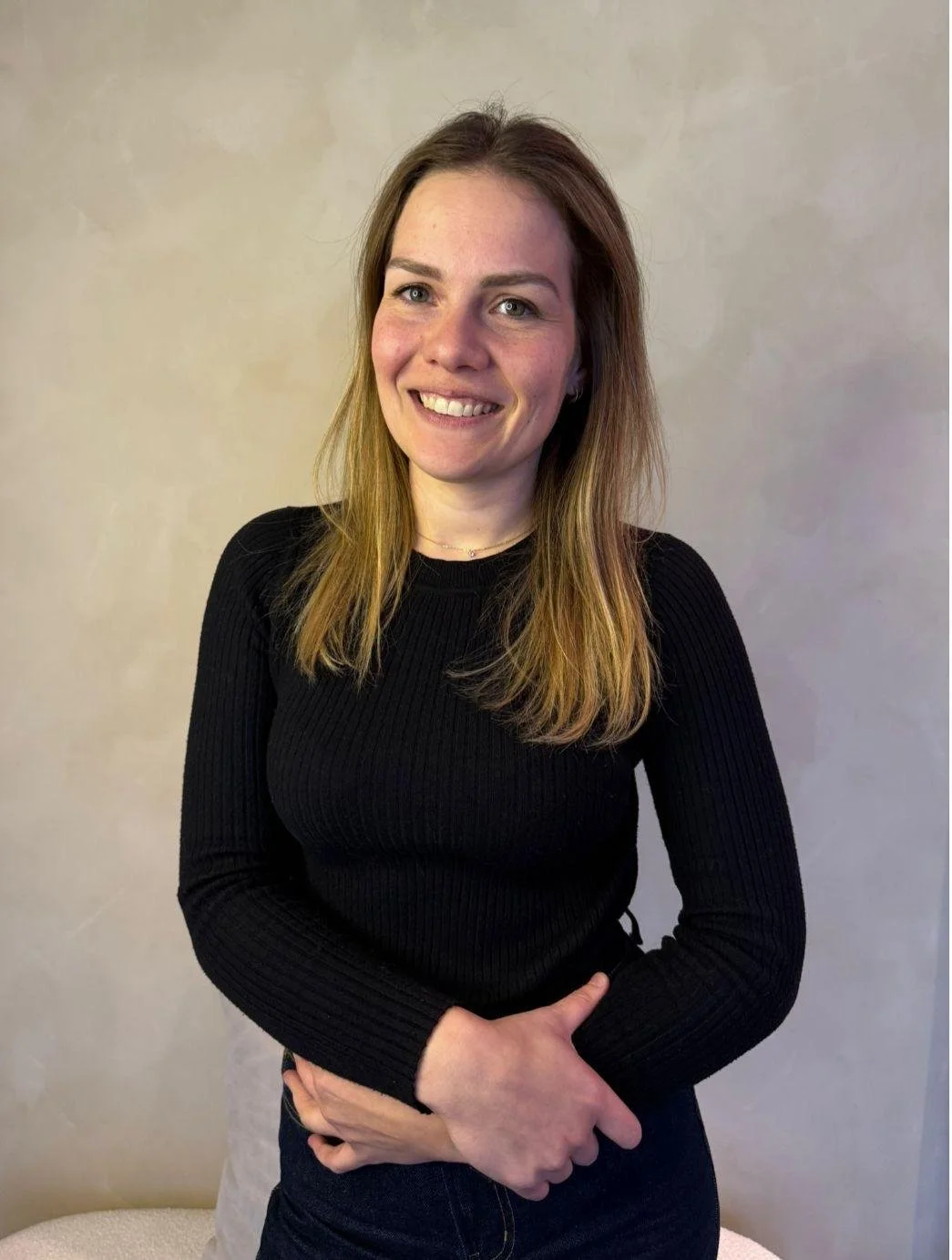 A man with curly dark hair, glasses, and a nose piercing, wearing a dark sweater, looking to the side with a slight smile, in a bright room with a plant in the background.