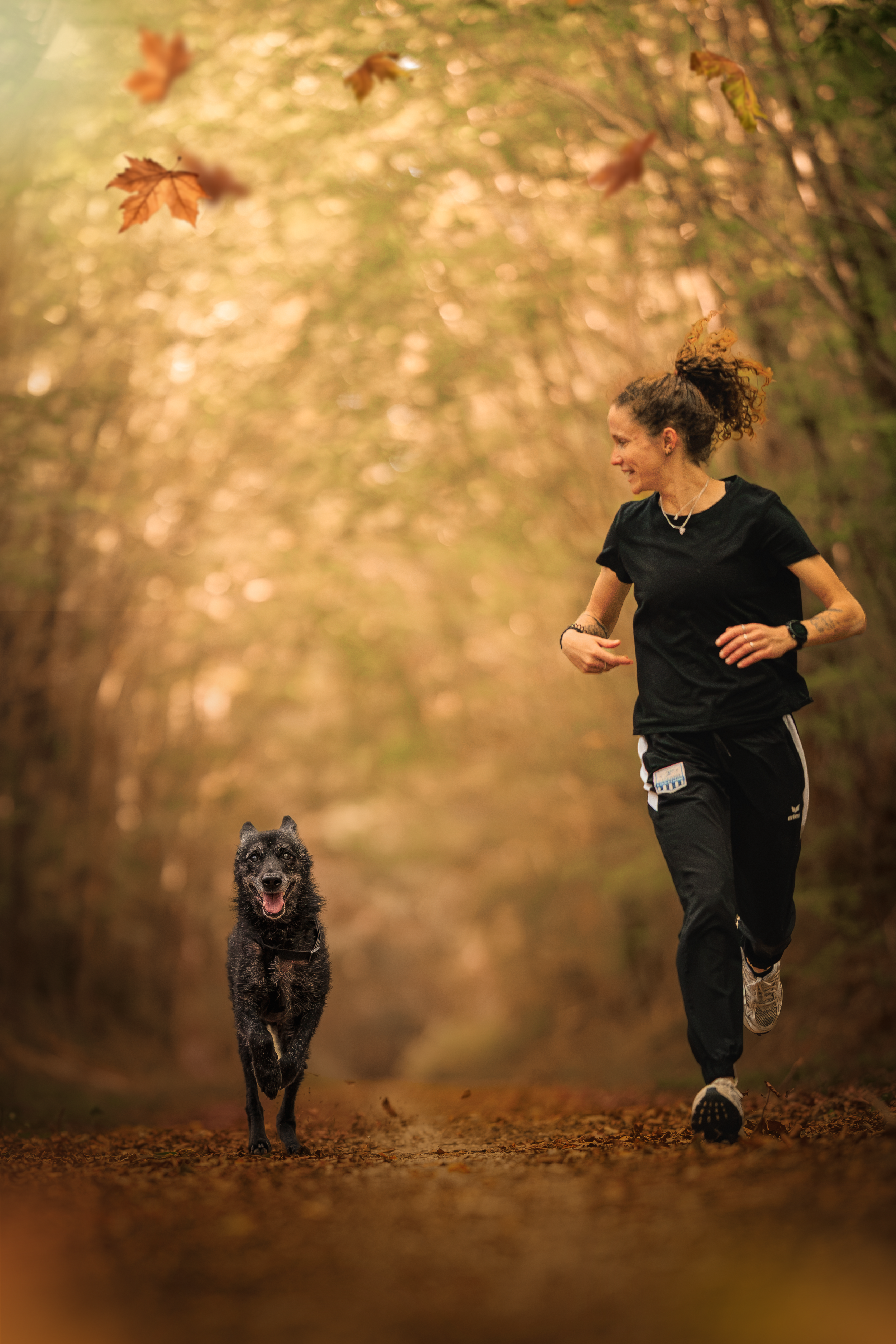 Une femme qui court dans un sentier forestier automnal avec un chien noir et feu. La femme porte un t-shirt noir, des pantalons de course, un bracelet et un collier, et a les cheveux attachés en un chignon. Le chien a l'air heureux, avec la langue sortie.