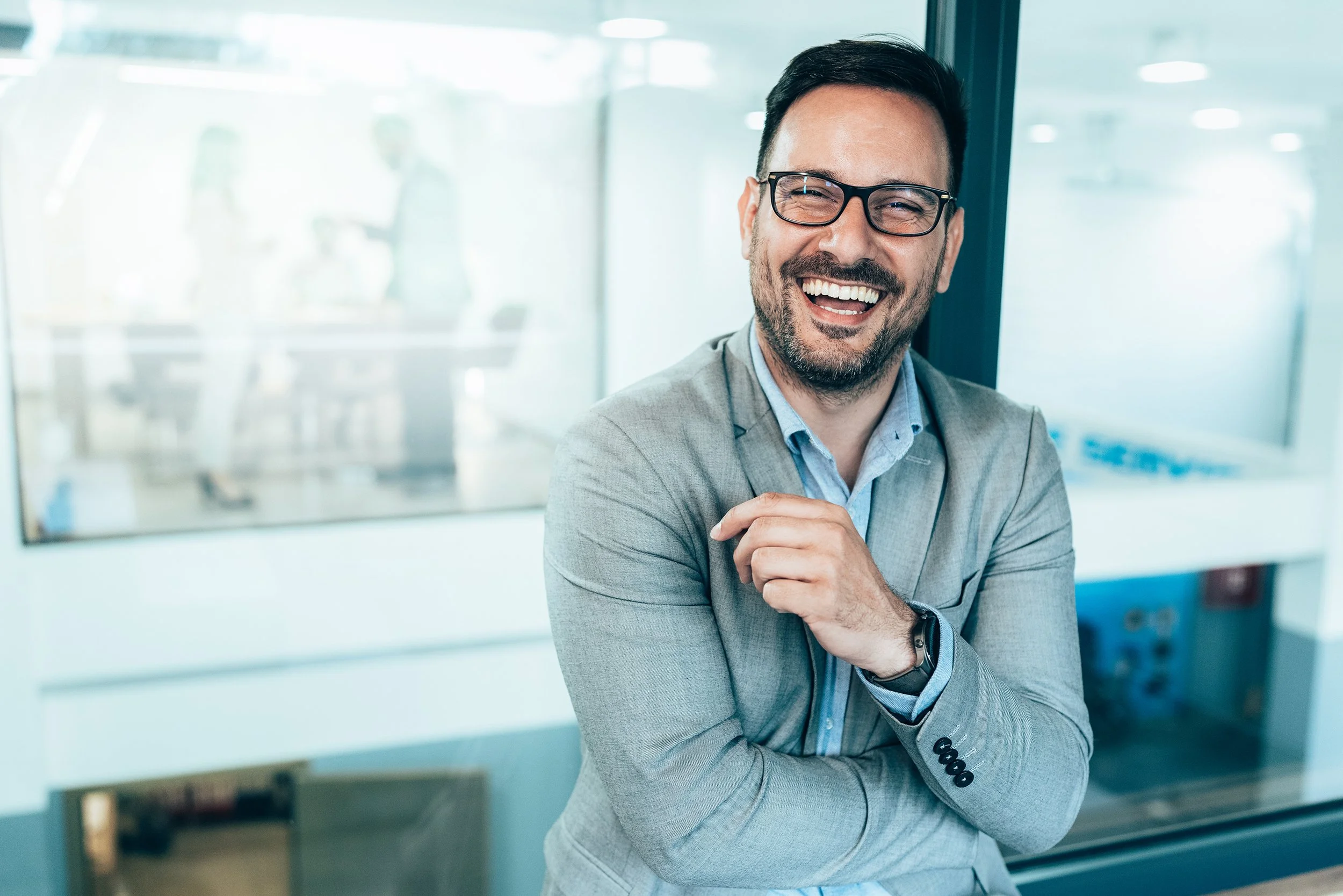A man in a gray suit and glasses smiling and laughing while standing in an office with glass walls.