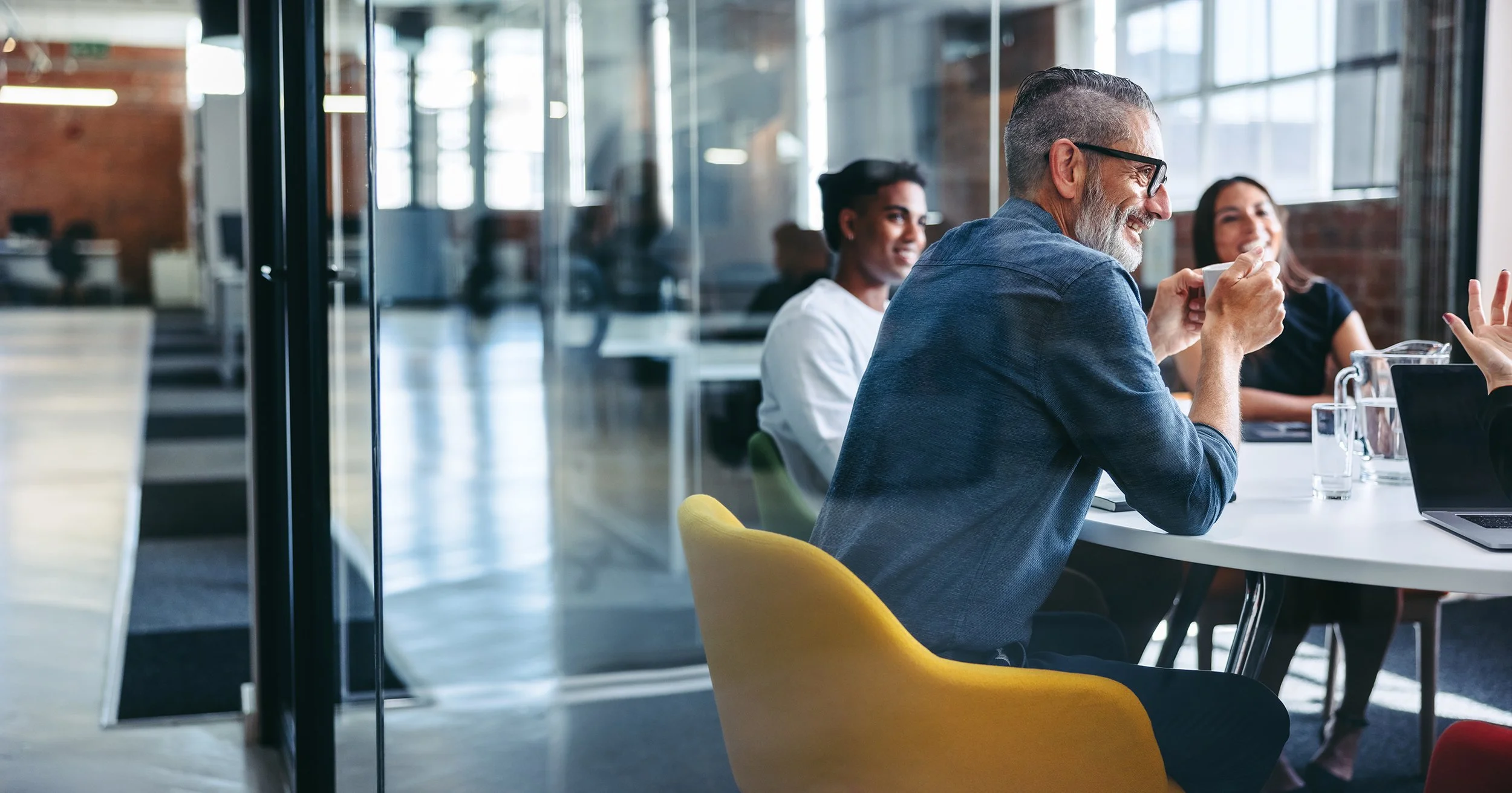 A group of professionals, including an older man with glasses, sitting together in a modern office or coworking space, engaging in a meeting or discussion, with one person sipping from a mug and others smiling, with large windows letting in natural light.