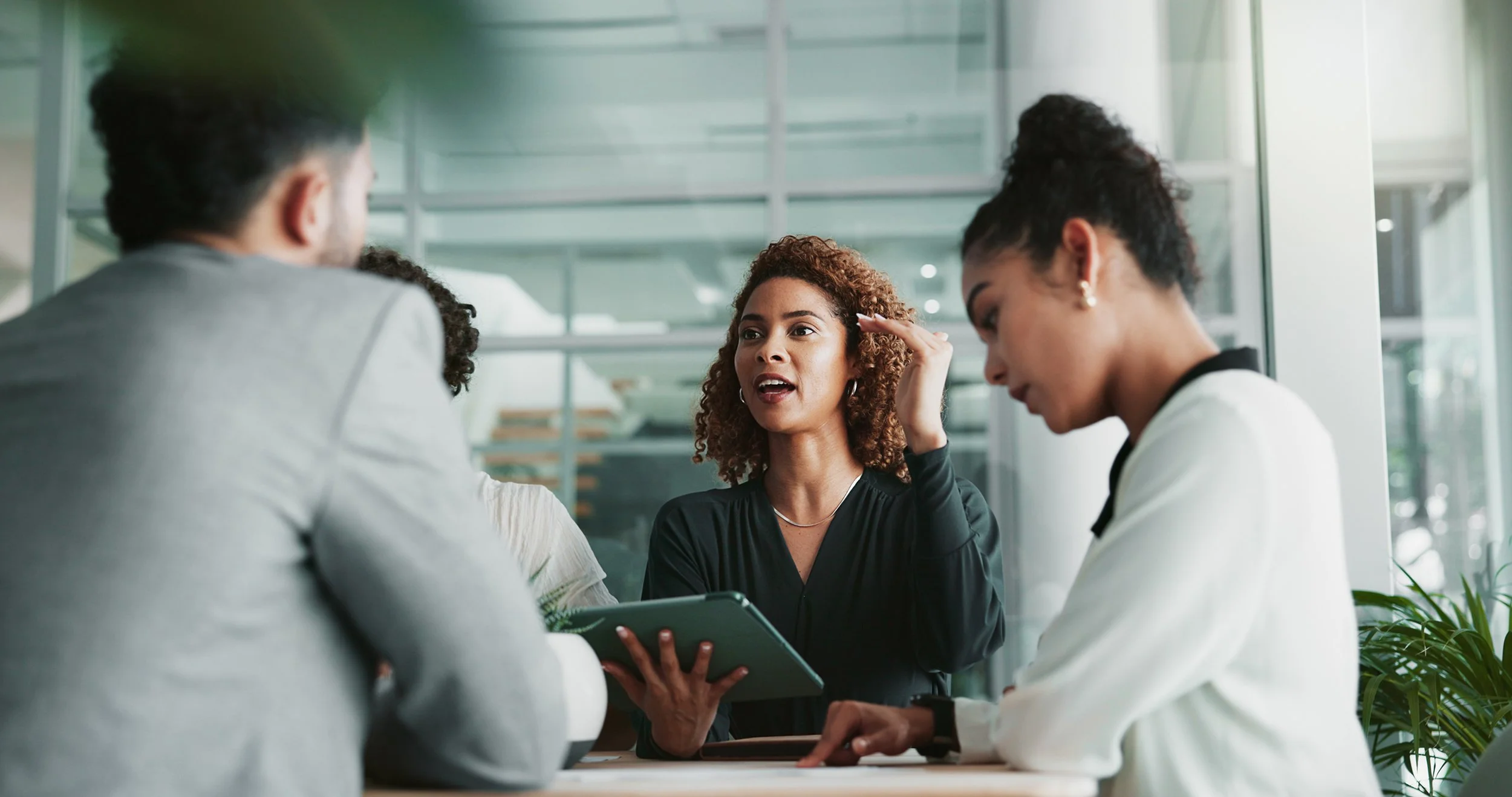Group of four diverse professionals having a discussion in a modern office, with one woman speaking and gesturing, others listening and taking notes.