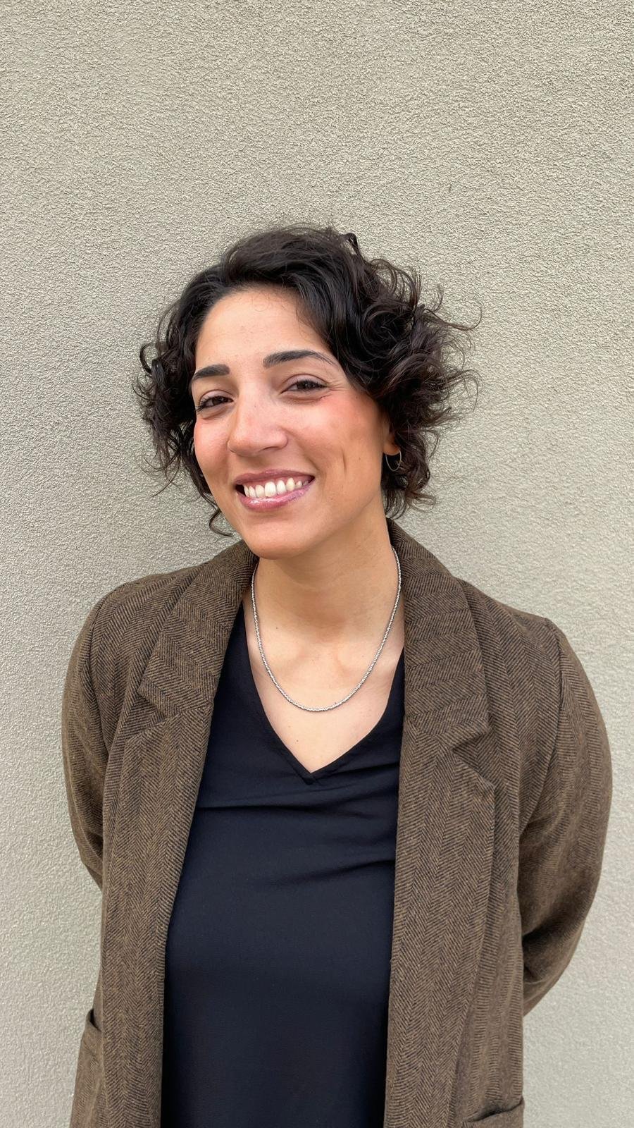 Ragazza sorridente con capelli ricci corti, indossa una giacca marrone, una maglietta nera e una collana, in piedi davanti a un muro beige.