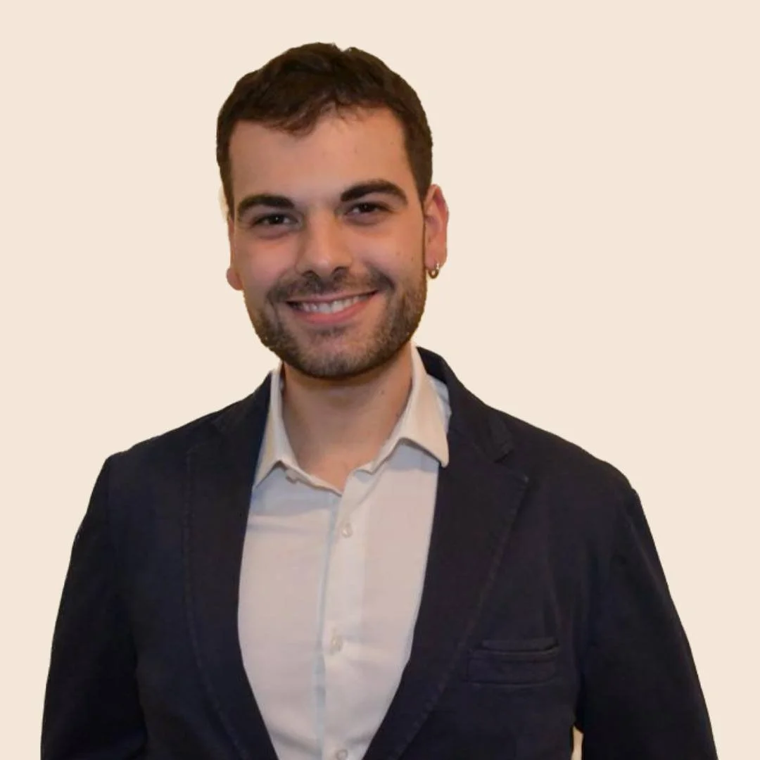 Uomo sorridente con capelli castani, barba curata, e orecchino, indossa una camicia bianca e una giacca nera, sfondo chiaro.