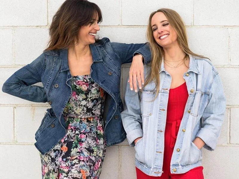 Two women smiling and chatting against a white brick wall, one in a floral dress with a denim jacket, and the other in a red dress with a light denim jacket.