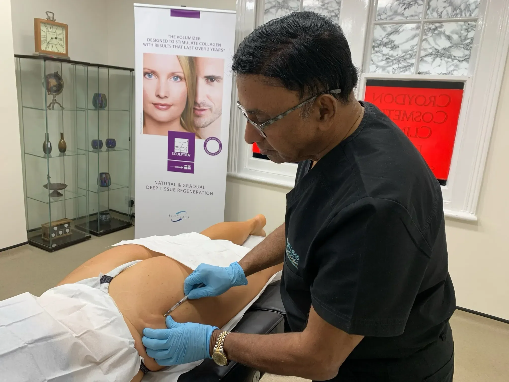 A medical professional in gloves administering an injection into a patient's abdomen in a clinical setting, with informational posters and display cabinets in the background.
