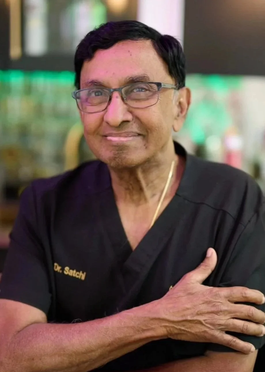 A man with black hair, glasses, and dark skin, smiling and wearing a black shirt with gold text, standing indoors with a blurred background.