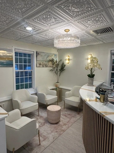 A stylish waiting area with four white chairs, a small round side table, and a curved reception desk. The room features a decorative tin ceiling, a chandelier, wall-mounted lights, and potted plants, with windows and wall art.
