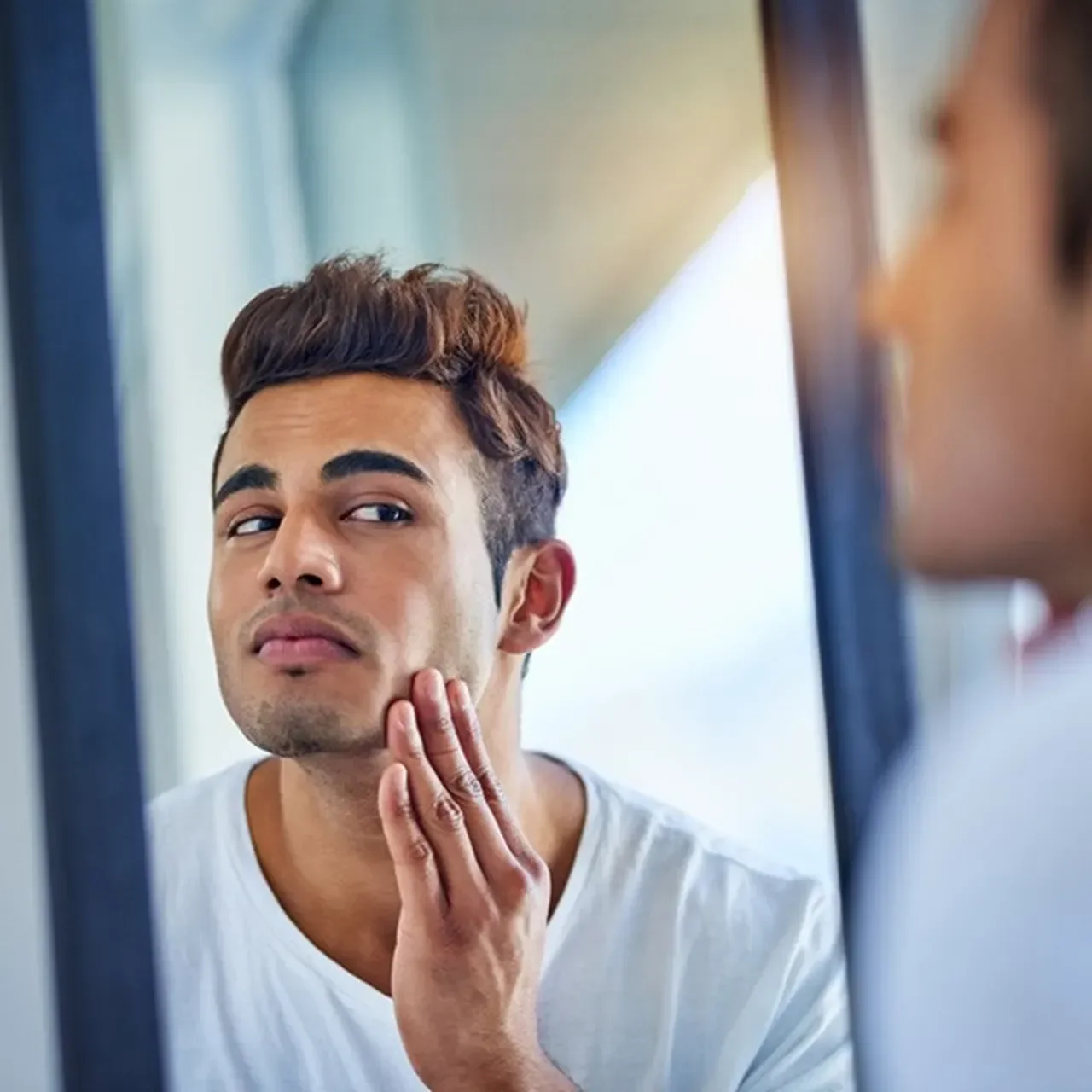 Young man examining his jawline in a mirror, touching his chin with his hand, with a concerned expression.