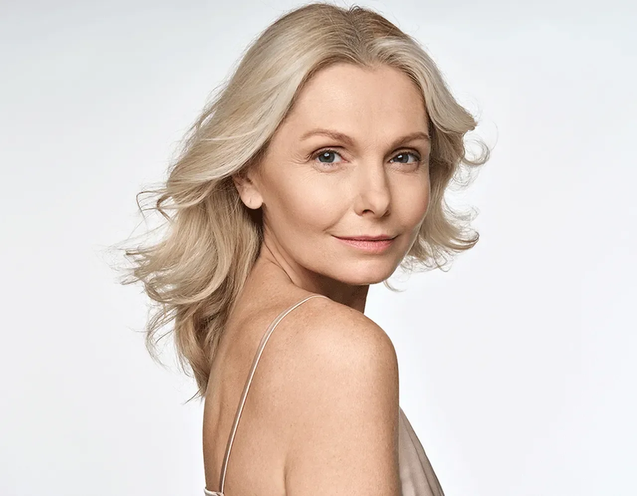 A portrait of a blonde woman with wavy hair, wearing a light-colored spaghetti strap top, smiling gently against a plain white background.