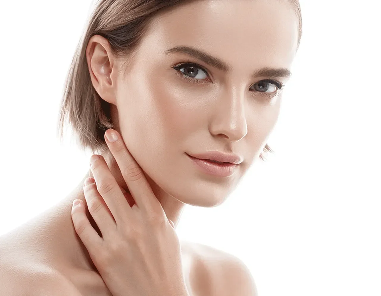 Close-up of a young woman with short brown hair, lightly touching her neck, with smooth skin and a gentle smile, against a white background.