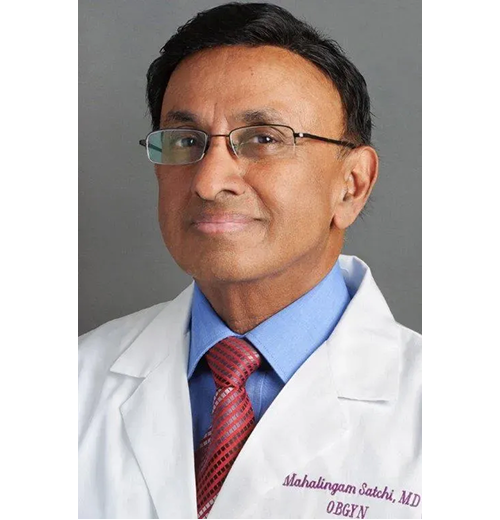 A professional portrait of a male doctor with glasses, wearing a white lab coat, blue shirt, and red tie, against a gray background.