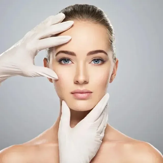 A woman with clear skin and blue eyes, posing with her hands wearing white gloves gently touching her face and neck against a light gray background.