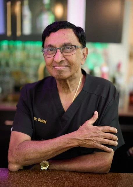 A man with glasses and dark hair, wearing a black medical uniform with gold embroidery, sitting at a bar or restaurant counter with his arms crossed and smiling.