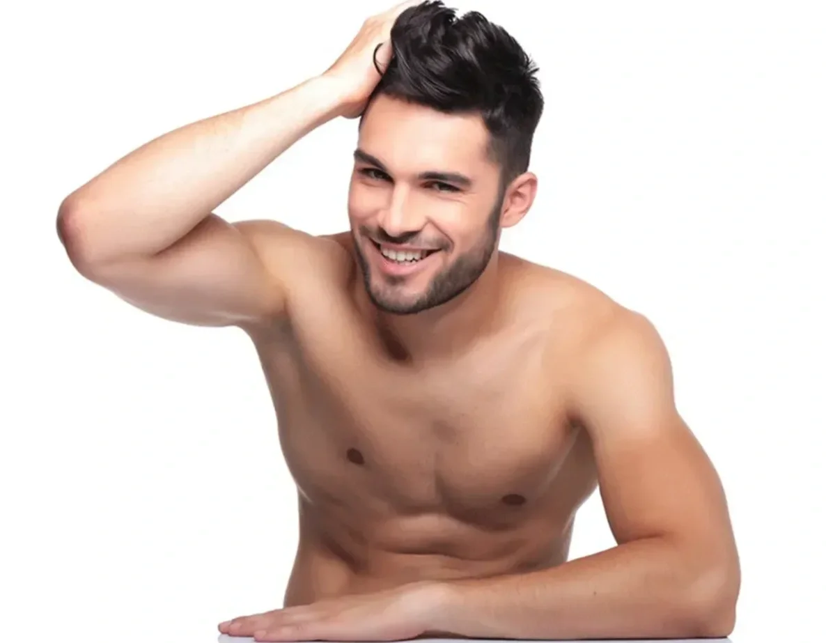 A shirtless man with short dark hair and a beard smiling, touching his head with one hand, against a white background.