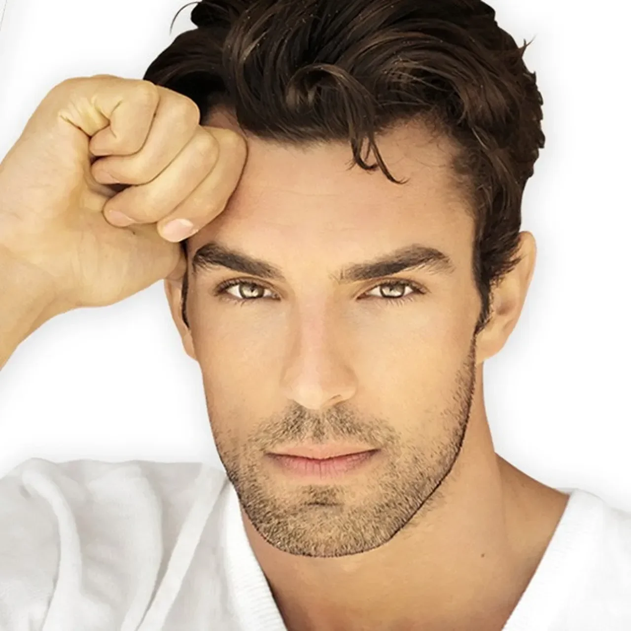A young man with dark, wavy hair and light eyes resting his head on his hand, looking directly at the camera against a white background.