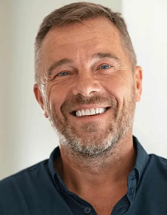 Close-up of a smiling middle-aged man with a beard and blue eyes, wearing a dark blue shirt, standing against a neutral background.