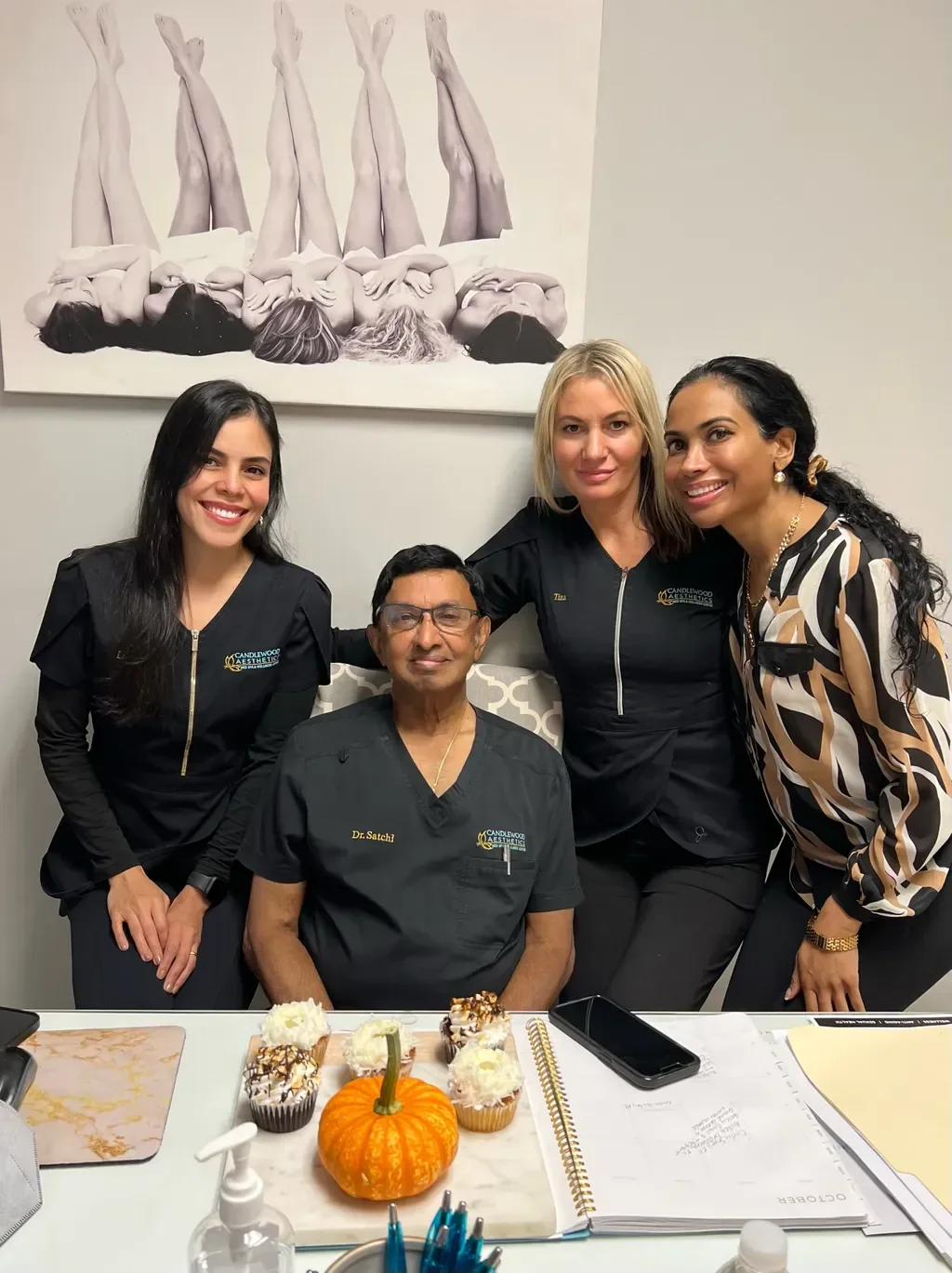 A group of four women and one man in a medical office, with a Halloween-themed cake and cupcakes on the table in front of them, and a black-and-white photograph of three women with their legs raised on the wall behind them.