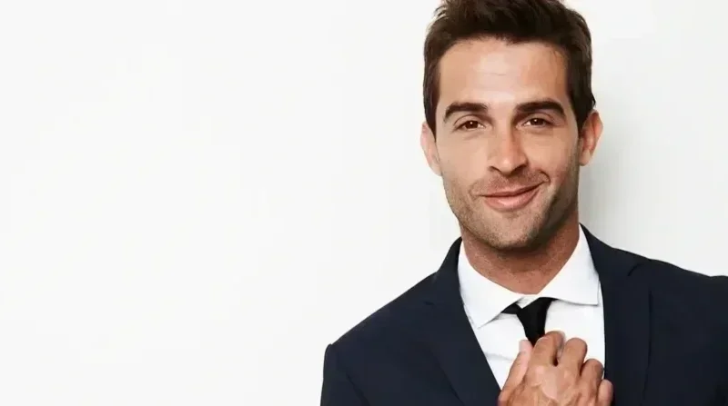 A man wearing a dark suit, white shirt, and black tie, standing against a white background, smiling confidently with one hand adjusting his tie.