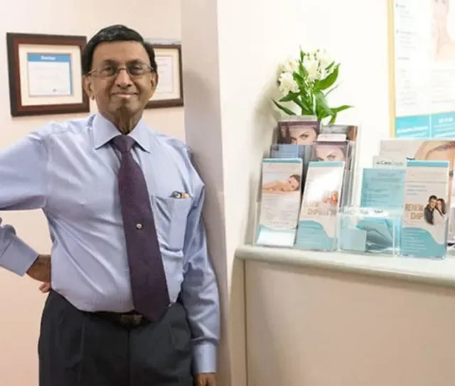 A man in a blue dress shirt and purple tie standing next to a countertop with brochures and pamphlets, with a small bouquet of white flowers on the counter, and framed certificates on the wall behind him.