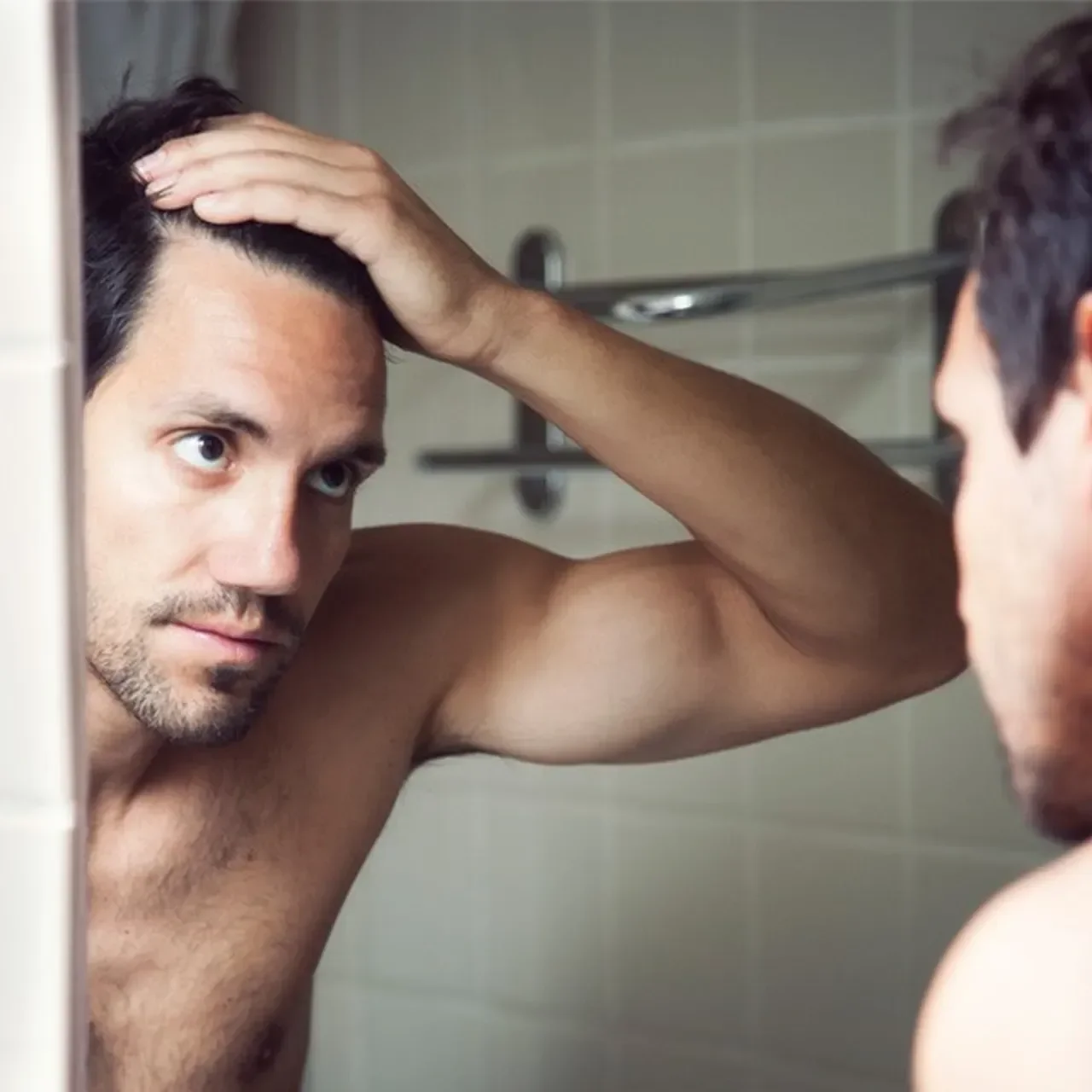 A man looking at himself in the mirror, touching his head, in a bathroom.