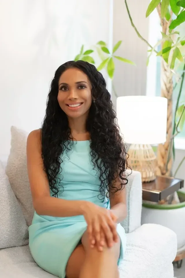 A woman with long, dark, curly hair sitting on a light-colored couch in a bright room, smiling at the camera, wearing a light blue sleeveless dress, with green plants and a table lamp in the background.