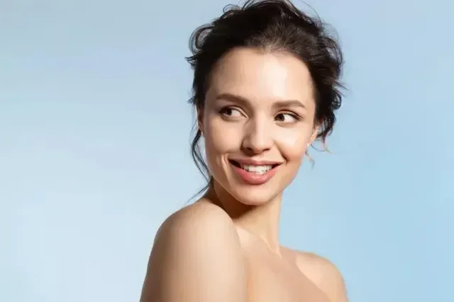 A woman with dark hair looking over her shoulder and smiling, against a light blue background.