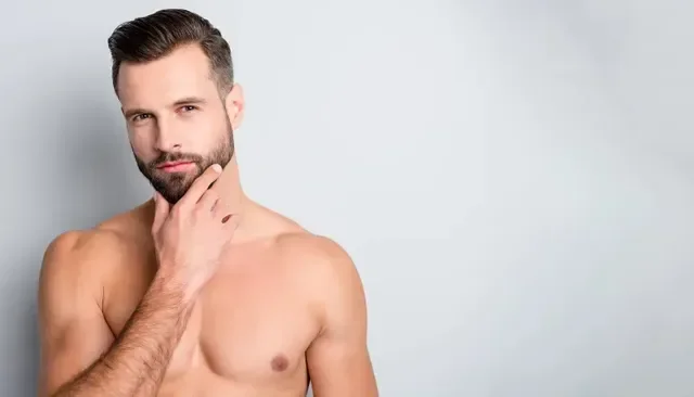 A shirtless man with a beard and short hair, touching his chin with his right hand, standing against a plain light gray background.