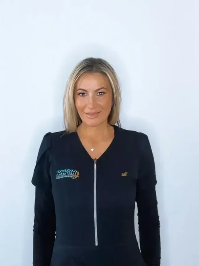A woman with blonde hair wearing a black zip-up jacket standing against a plain white wall.