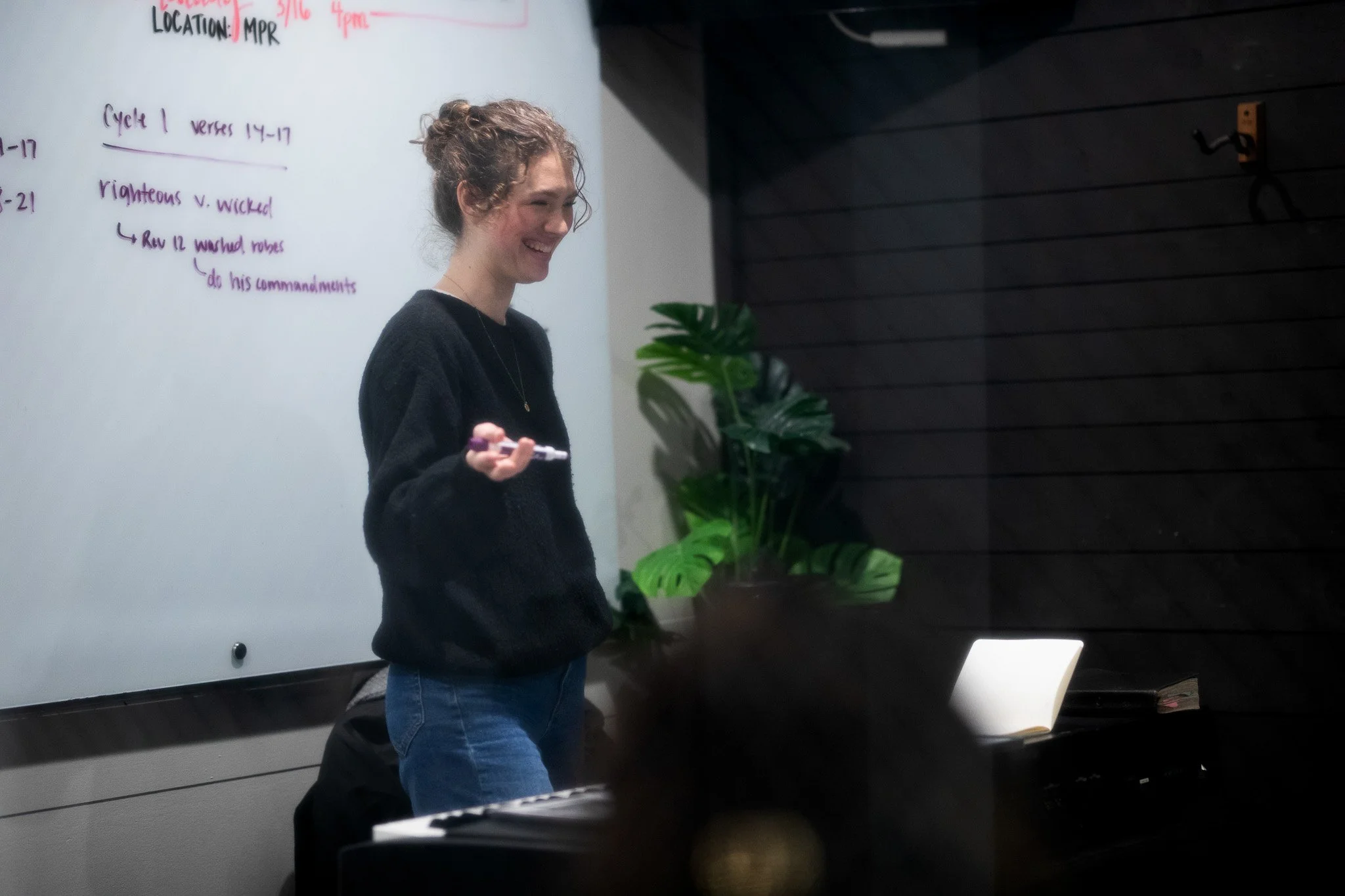 A young woman with curly hair, smiling, holding a marker, in a classroom or meeting room with a whiteboard, notes, and green houseplant.