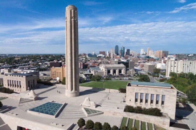 Aerial view of a cityscape with a tall monument, surrounded by historic buildings, greenery, and modern skyscrapers in the background.