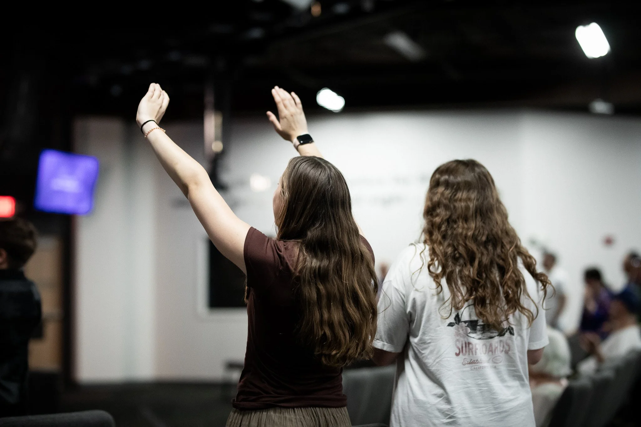 Two women with long hair, one with a black wristwatch, raising their hands in an indoor setting with a large presentation screen and other people in the background.