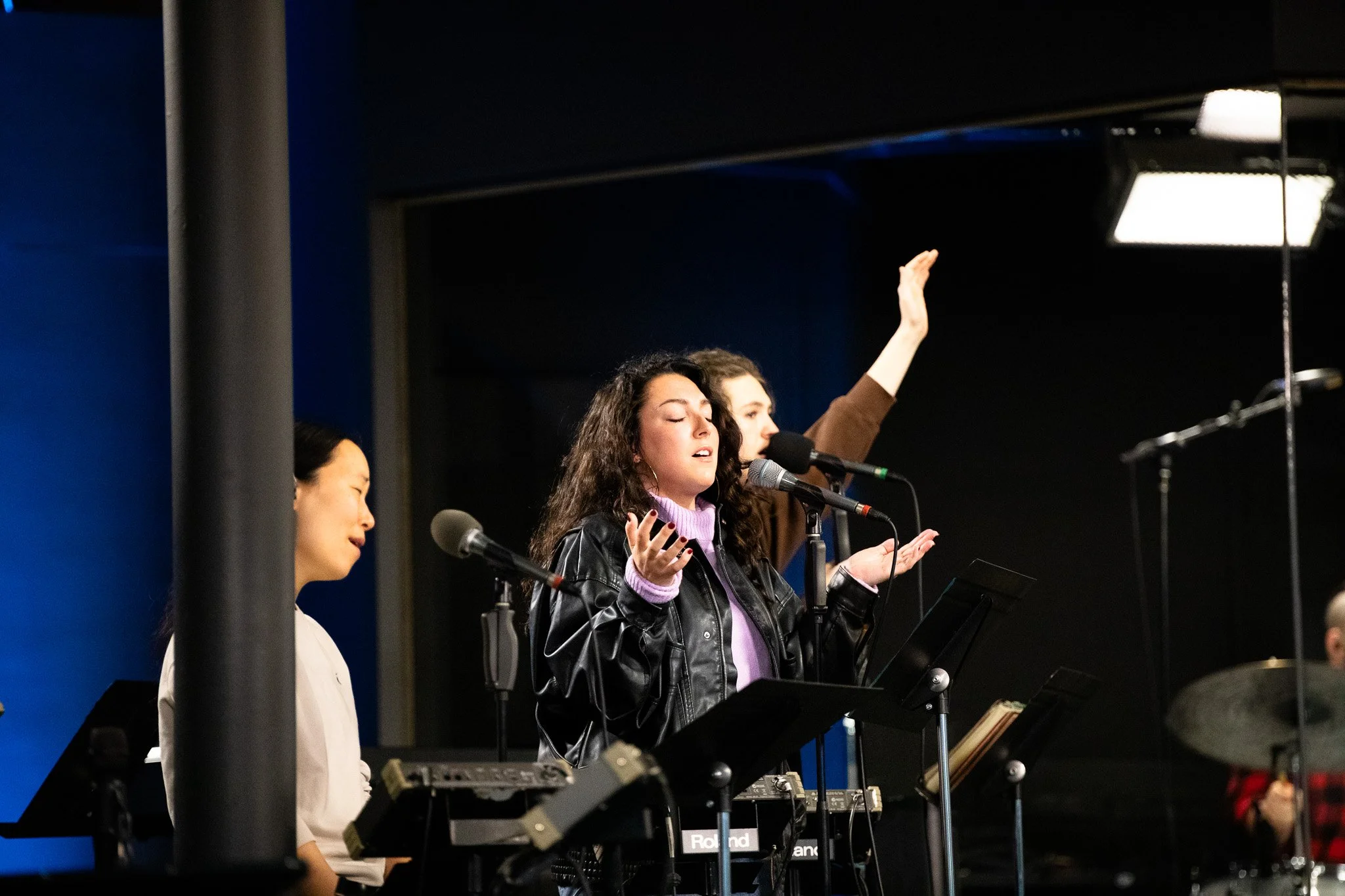A group of women singing and playing musical instruments in a recording studio, with one woman raising her hand and singing into a microphone.