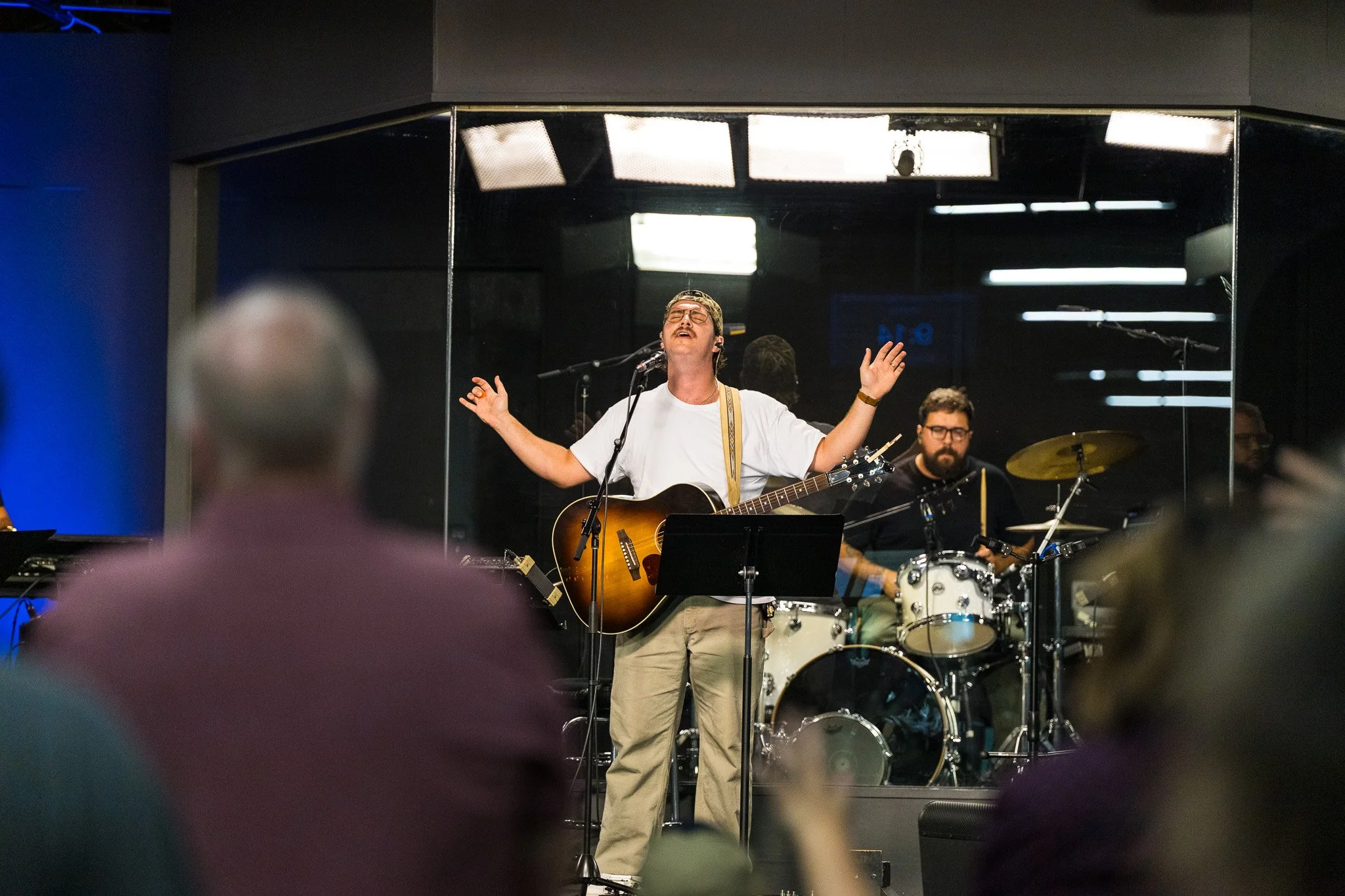 A man singing passionately with eyes closed while playing guitar during a live music performance, accompanied by other musicians, in a dimly lit venue with stage lights and a reflective glass backdrop.