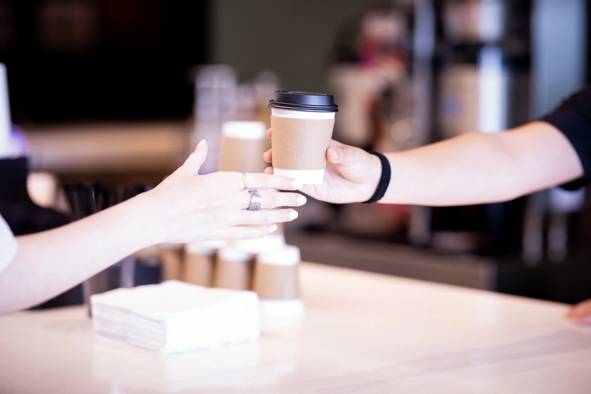 A person handing a takeaway coffee cup with a cardboard sleeve to another person at a cafe.