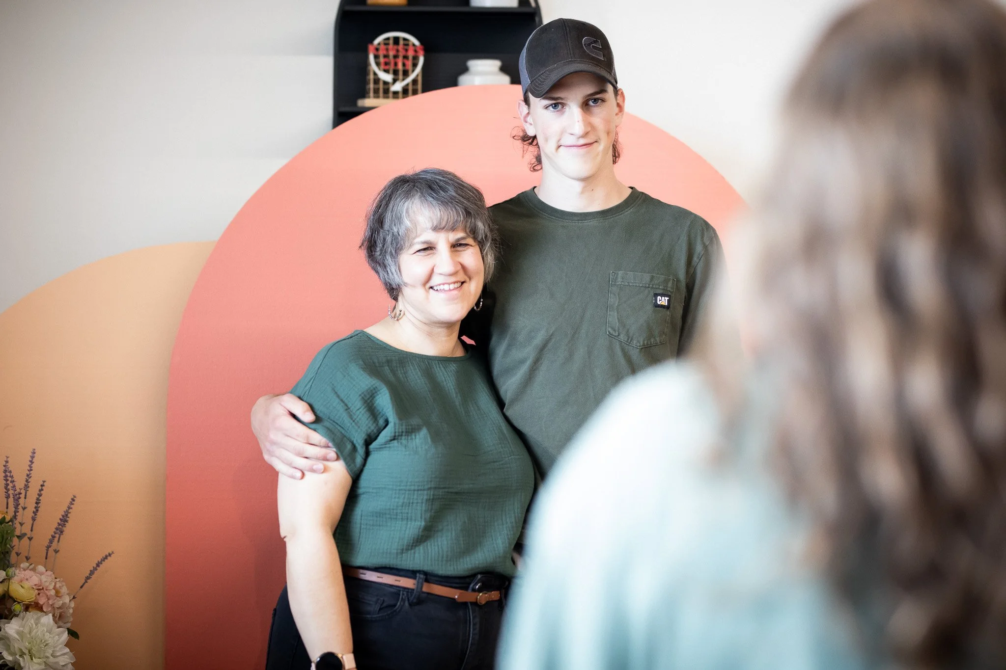A woman and a young man are standing together, smiling for a photo. The woman has gray hair and is wearing a green top, while the young man has curly hair, wearing a black baseball cap and a green shirt with a CAT logo. They are indoors with decorative elements in the background and a person in the foreground partially visible.