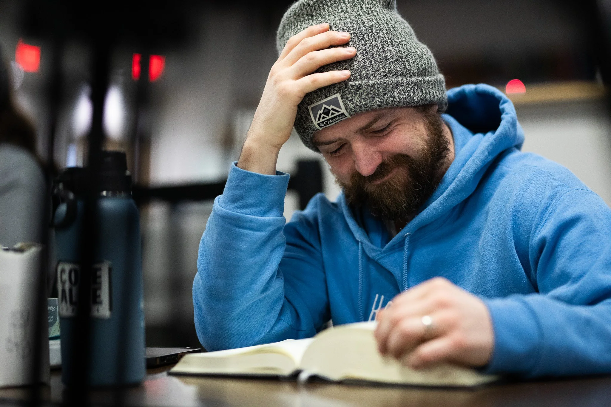A man with a beard wearing a gray knit beanie and a blue hoodie, holding his head with one hand and sitting at a table with an open notebook in front of him. The background is blurry with a dark setting and some red lights.