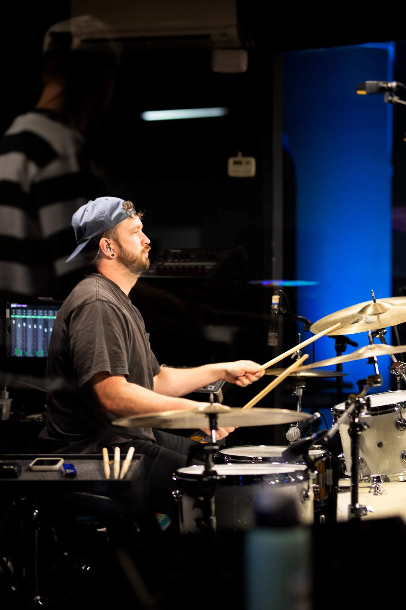 A male drummer wearing a backward cap and a dark t-shirt playing drums in a dimly lit studio or stage setting.