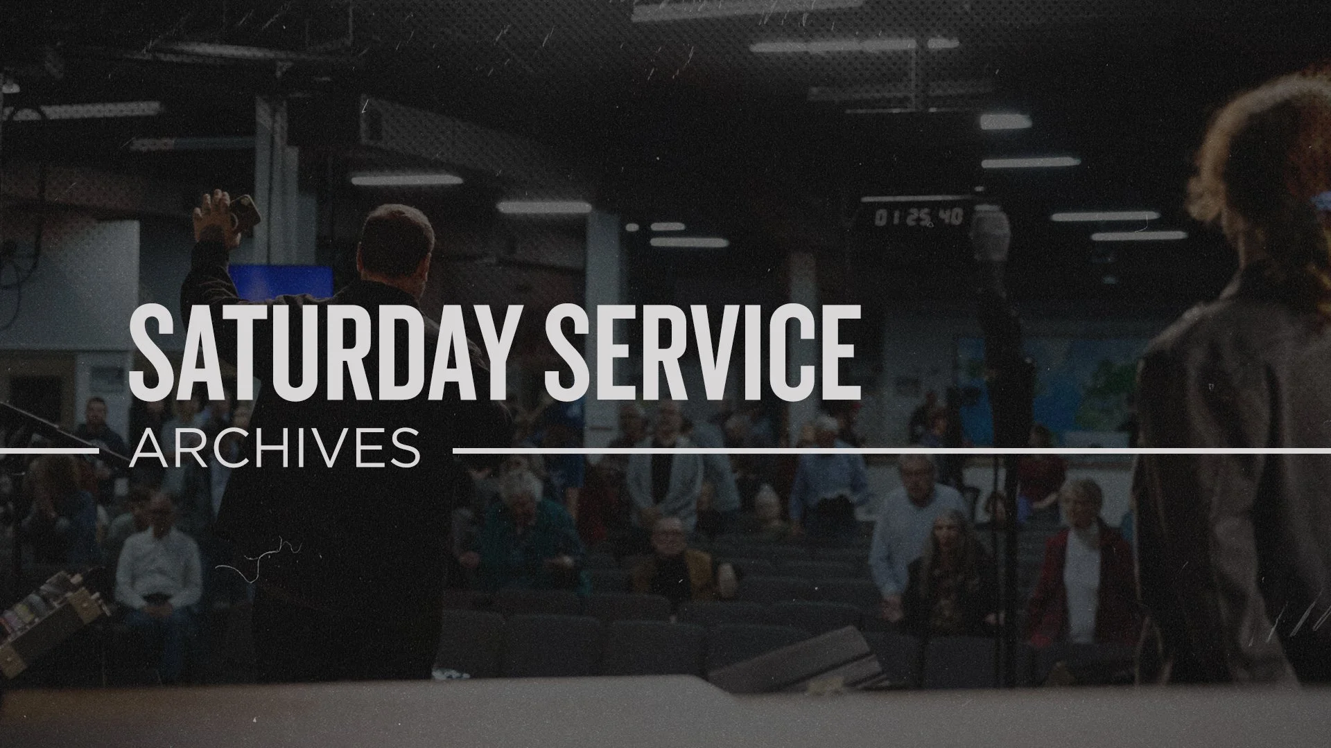 A group of people gathered indoors, with some standing and others seated, watching a presentation or event. The scene is slightly blurred and dark, with large text overlay that reads 'Saturday Service Archives'.